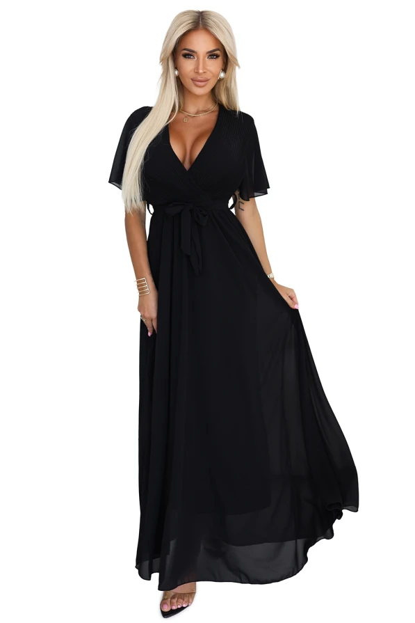 Modelka prezentuje elegancką czarną sukienkę maxi z szyfonu, z plisowanym dekoltem w kształcie litery V, krótkimi rękawkami i wiązaniem w talii; lekka i kobieca stylizacja marki Numoco Basic, idealna na wyjątkowe okazje.