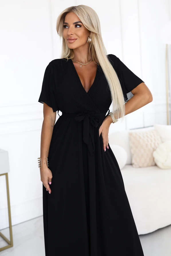 Modelka prezentuje elegancką czarną sukienkę maxi z szyfonu, z plisowanym dekoltem w kształcie litery V, krótkimi rękawkami i wiązaniem w talii; lekka i kobieca stylizacja marki Numoco Basic, idealna na wyjątkowe okazje.