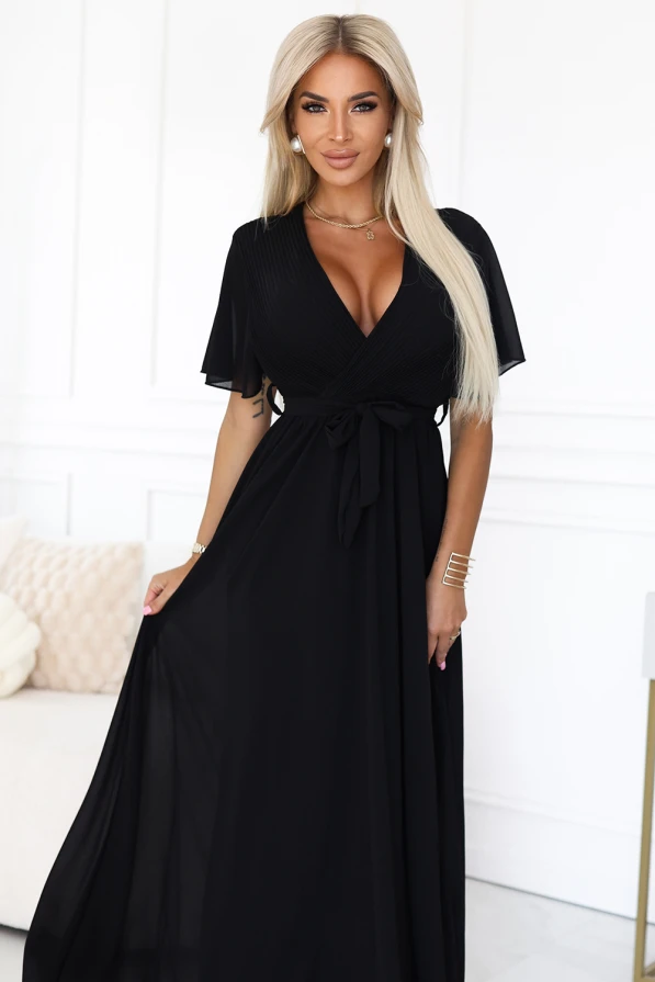 Modelka prezentuje elegancką czarną sukienkę maxi z szyfonu, z plisowanym dekoltem w kształcie litery V, krótkimi rękawkami i wiązaniem w talii; lekka i kobieca stylizacja marki Numoco Basic, idealna na wyjątkowe okazje.