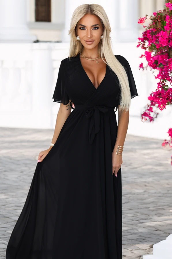 Modelka prezentuje elegancką czarną sukienkę maxi z szyfonu, z plisowanym dekoltem w kształcie litery V, krótkimi rękawkami i wiązaniem w talii; lekka i kobieca stylizacja marki Numoco Basic, idealna na wyjątkowe okazje.