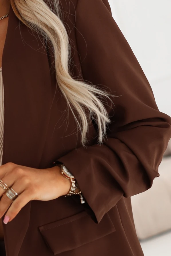 Czekoladowa marynarka damska o przedłużanym kroju z klapami. Modelka w brązowym żakiecie w stylizacji casual elegance.