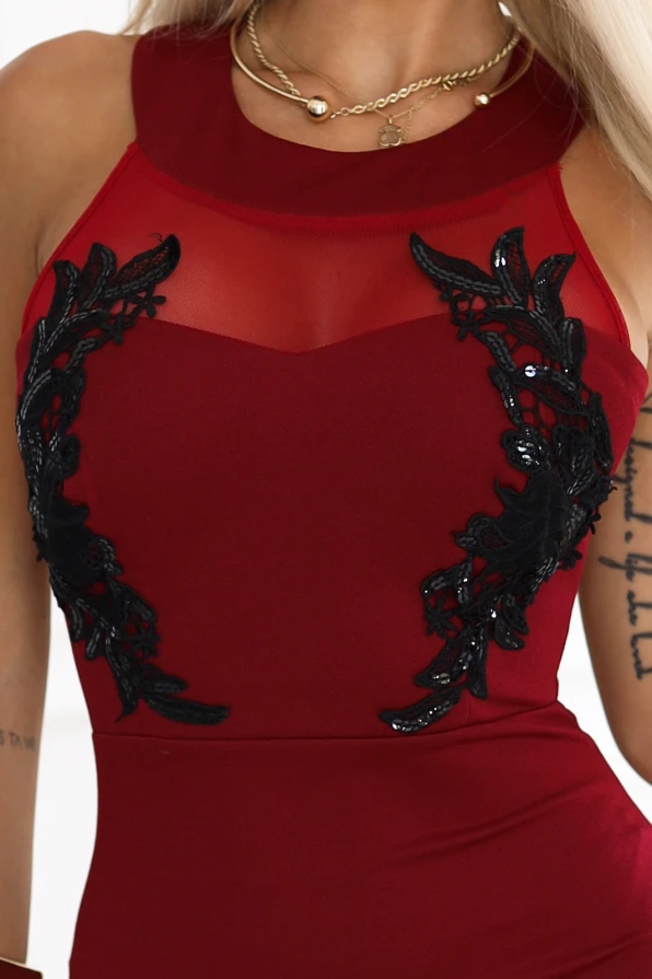 Modelka w długiej bordowej sukni wieczorowej z koronkowymi zdobieniami i siateczką przy dekolcie, stojąca w eleganckim, jasnym wnętrzu. Sukienka podkreśla talię i eksponuje kobiecą sylwetkę – idealna na sylwestra, wesele lub galę.