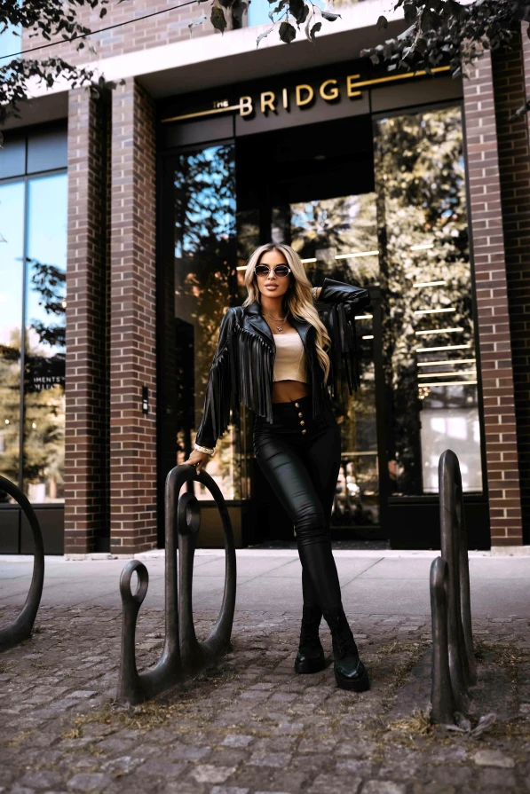 Modelka prezentuje czarną ramoneskę z frędzlami marki Numoco Basic, tworząc stylową i nowoczesną stylizację w miejskim otoczeniu.