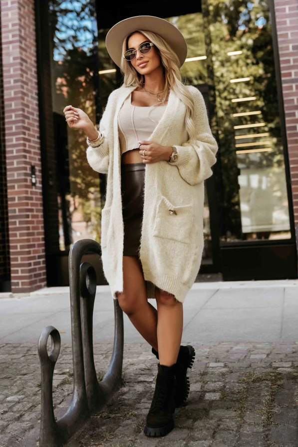 Modelka prezentuje długi kardigan damski z miękkiej dzianiny w kolorze beżowym, z kieszeniami i ozdobną broszką – Numoco Basic.