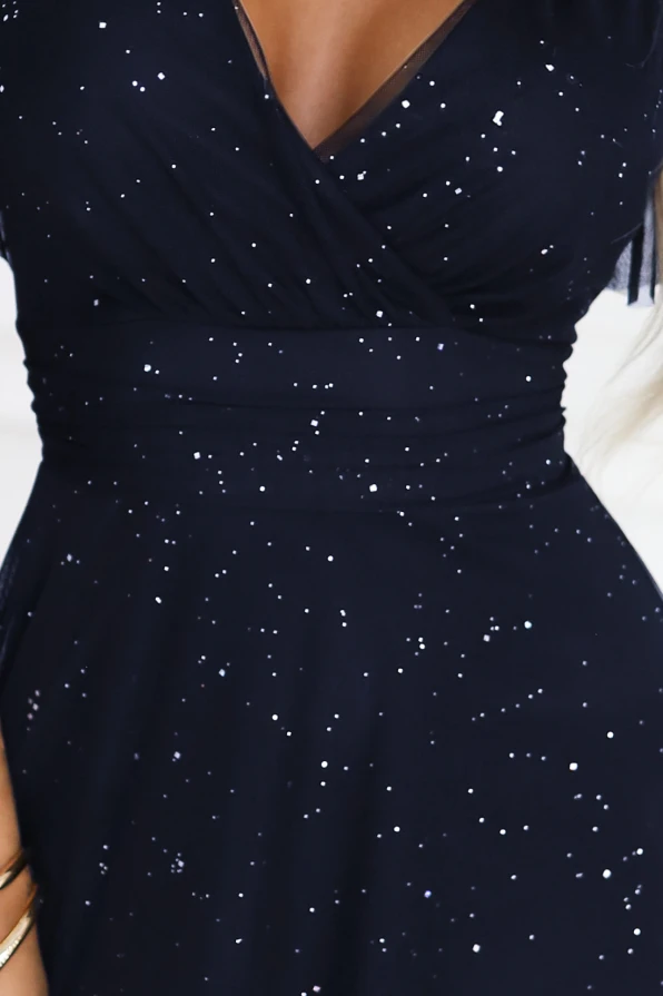 „Modelka prezentuje długą, granatową suknię wieczorową Numoco z połyskującymi drobinkami, kopertowym dekoltem i zwiewnymi rękawkami, stojąc w eleganckim, jasnym wnętrzu.