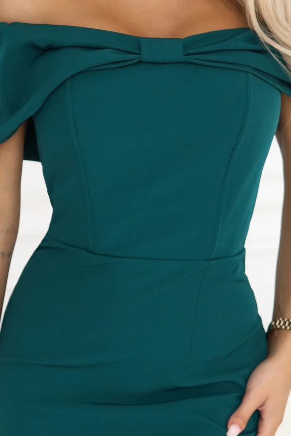 Wieczorowa sukienka maxi w odcieniu butelkowej zieleni z kokardą na dekolcie, pokazana na modelce w eleganckiej aranżacji.